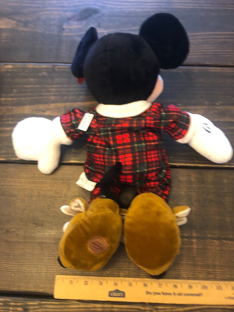 May include: A plush Mickey Mouse toy wearing a red and green plaid shirt and brown shoes. The toy is lying on its back with its arms outstretched.