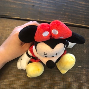 Op de afbeelding: Een pluche speelgoed van Minnie Mouse met een rode strik, zwarte oren en gele schoenen. Het speelgoed zit op een houten oppervlak.