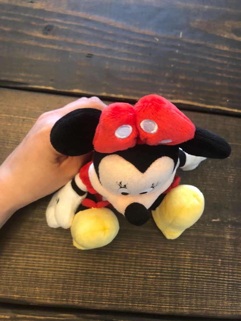 Op de afbeelding: Een pluche speelgoed van Minnie Mouse met een rode strik, zwarte oren en gele schoenen. Het speelgoed zit op een houten oppervlak.