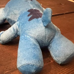 Op de afbeelding: Een lichtblauw pluche speelgoed met een donkerblauwe vlek op de rug. Het speelgoed heeft een zachte, fuzzy textuur en ligt op een houten oppervlak. Het speelgoed heeft een staart en vier ledematen.
