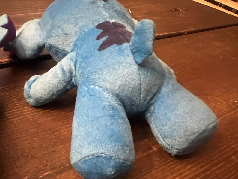 Op de afbeelding: Een lichtblauw pluche speelgoed met een donkerblauwe vlek op de rug. Het speelgoed heeft een zachte, fuzzy textuur en ligt op een houten oppervlak. Het speelgoed heeft een staart en vier ledematen.