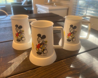 May include: Three white plastic mugs featuring Mickey Mouse playing a guitar. The mugs have a flared base and a rectangular handle. The Mickey Mouse graphic is in color, with a red shirt and yellow accents. The mugs are on a dark wooden table.