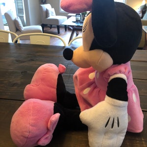 May include: A pink and black plush toy of Minnie Mouse. She is wearing a pink dress with white polka dots and a pink bow. She has a white glove on her right hand.