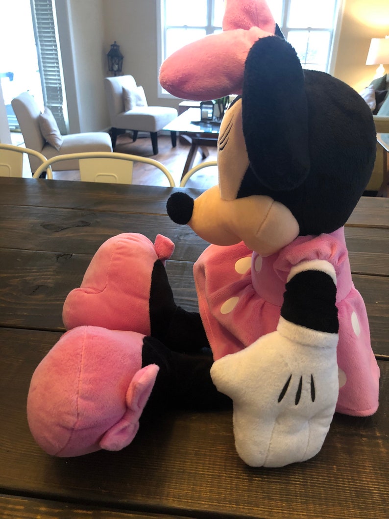 May include: A pink and black plush toy of Minnie Mouse. She is wearing a pink dress with white polka dots and a pink bow. She has a white glove on her right hand.