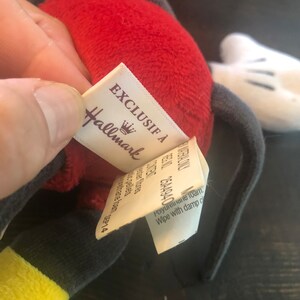 Puede incluir: Un juguete de peluche rojo y gris con un guante blanco. El juguete tiene una etiqueta que dice "Exclusif &agrave; Hallmark" con un logotipo de corona.