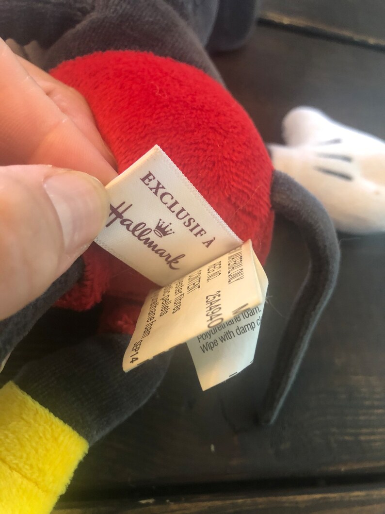 Puede incluir: Un juguete de peluche rojo y gris con un guante blanco. El juguete tiene una etiqueta que dice "Exclusif &agrave; Hallmark" con un logotipo de corona.