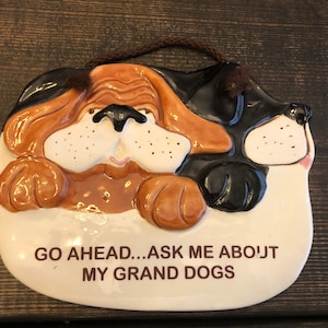 May include: A white ceramic plaque with a brown rope hanger. The plaque features a cartoon image of two dogs, one brown and one black and white, with the text "GO AHEAD...ASK ME ABOUT MY GRAND DOGS".