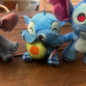 Op de afbeelding: Verschillende Stitch-knuffels in verschillende poses. E&eacute;n blauwe Stitch-knuffel knuffelt een avocado-knuffel. Een andere Stitch heeft een hartvormig oog. De knuffels staan op een houten oppervlak.