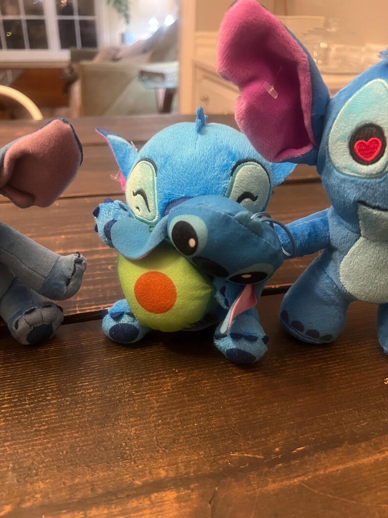 Op de afbeelding: Verschillende Stitch-knuffels in verschillende poses. E&eacute;n blauwe Stitch-knuffel knuffelt een avocado-knuffel. Een andere Stitch heeft een hartvormig oog. De knuffels staan op een houten oppervlak.