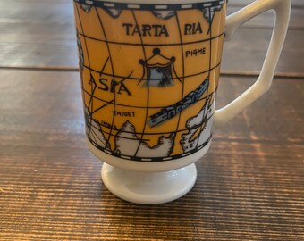 May include: White ceramic mug with a black and yellow world map design. The map features the text "TARTARIA" and "ASIA".