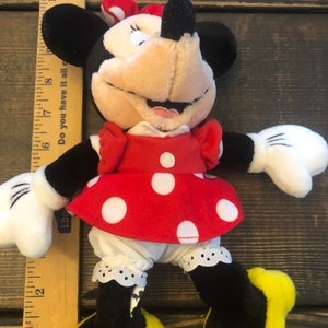 May include: A plush toy of Minnie Mouse wearing a red dress with white polka dots, a black bow, and yellow shoes. The toy is about 8 inches tall and is made of soft, cuddly material.