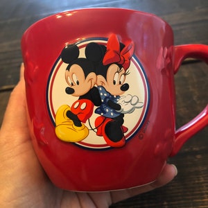 May include: Red ceramic mug with a circular design featuring Mickey Mouse and Minnie Mouse. The design is in a cartoon style and includes a red bow, yellow shoes, and blue polka dots. The mug has a handle and the text "Disney" is printed on the bottom right corner.