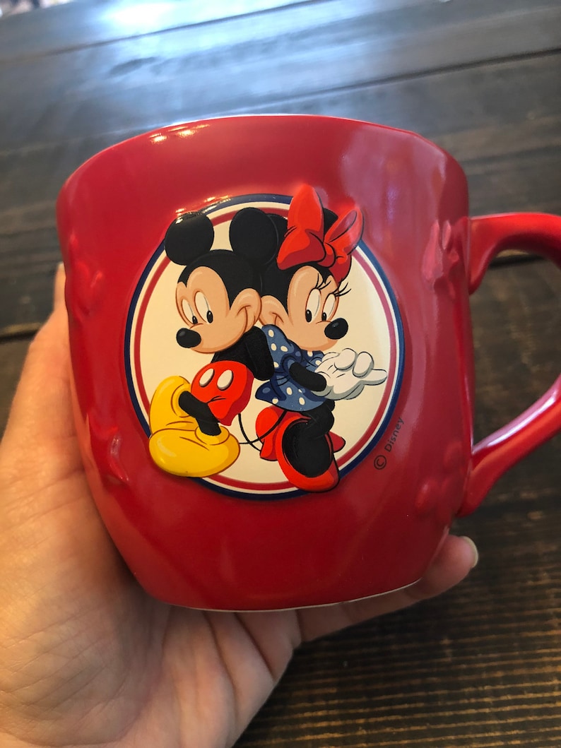 May include: Red ceramic mug with a circular design featuring Mickey Mouse and Minnie Mouse. The design is in a cartoon style and includes a red bow, yellow shoes, and blue polka dots. The mug has a handle and the text "Disney" is printed on the bottom right corner.