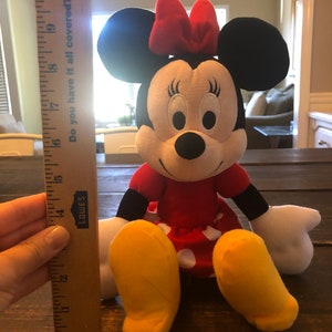 May include: A plush red and black Minnie Mouse toy with a red bow, white polka dot skirt, and yellow shoes. The toy is sitting on a wooden surface.