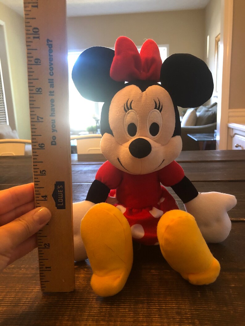 May include: A plush red and black Minnie Mouse toy with a red bow, white polka dot skirt, and yellow shoes. The toy is sitting on a wooden surface.