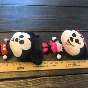 May include: Two plush Mickey Mouse toys. One is black with sparkly details and red shorts. The other is beige with black sparkly details and a pink dress. Both have black ears and a black nose.