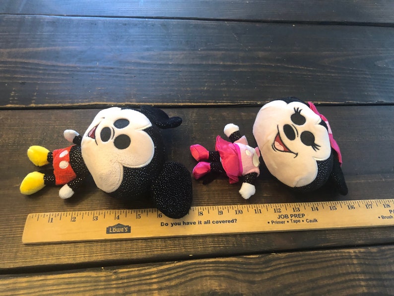 May include: Two plush Mickey Mouse toys. One is black with sparkly details and red shorts. The other is beige with black sparkly details and a pink dress. Both have black ears and a black nose.
