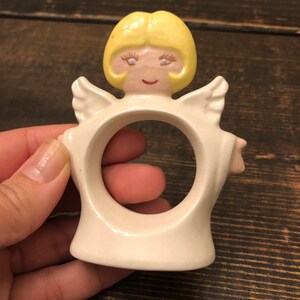 Puede incluir: Anillo de servilleta de cer&aacute;mica blanca con forma de &aacute;ngel con cabello amarillo, ojos azules y rostro rosado.