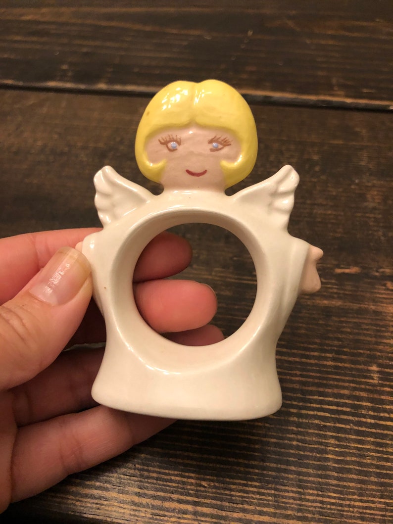 Puede incluir: Anillo de servilleta de cer&aacute;mica blanca con forma de &aacute;ngel con cabello amarillo, ojos azules y rostro rosado.