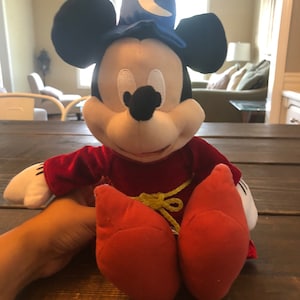 May include: A plush Mickey Mouse toy wearing a blue wizard hat with a yellow crescent moon. He is wearing a red velvet robe with a yellow rope belt and red shoes.