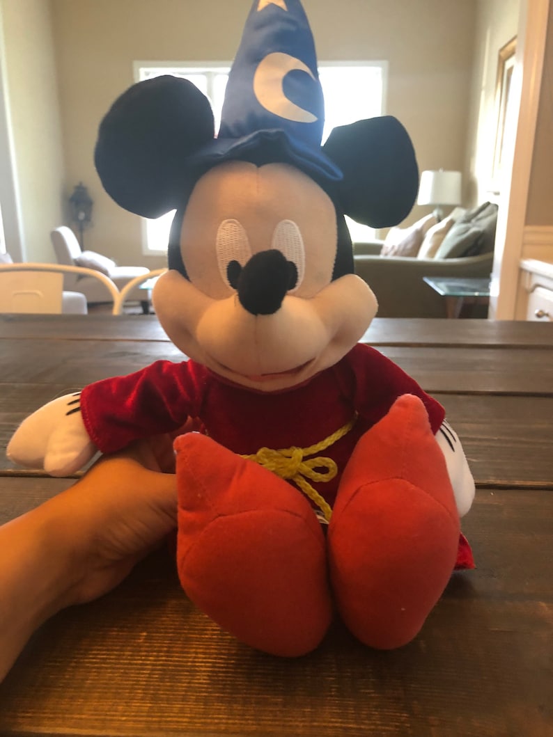May include: A plush Mickey Mouse toy wearing a blue wizard hat with a yellow crescent moon. He is wearing a red velvet robe with a yellow rope belt and red shoes.