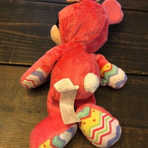 Op de afbeelding: Roze pluche konijn speelgoed met een witte staart en een kleurrijk chevron patroon op de benen. Het konijn draagt een roze en paarse paashazenhoed.