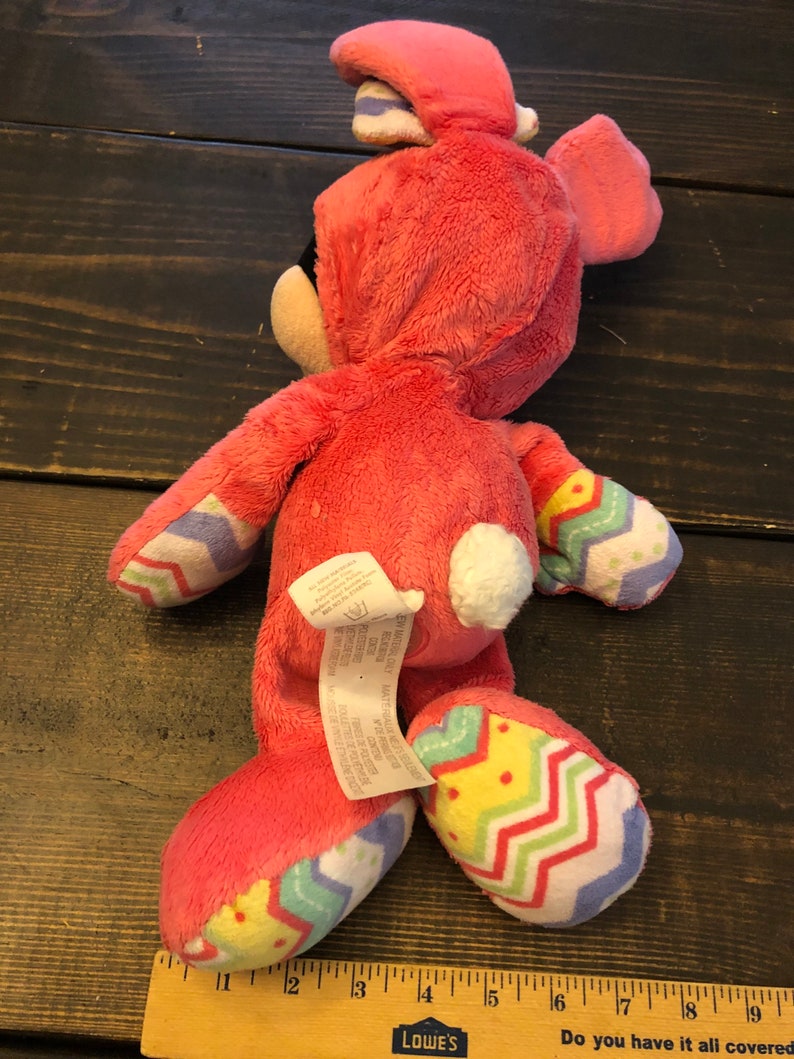 Op de afbeelding: Roze pluche konijn speelgoed met een witte staart en een kleurrijk chevron patroon op de benen. Het konijn draagt een roze en paarse paashazenhoed.