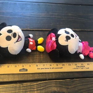May include: Two plush toys, Mickey Mouse and Minnie Mouse, are lying on a wooden surface. Mickey is black with red shorts and yellow shoes. Minnie is black with a pink bow and pink dress. Both toys have sparkly black fabric.