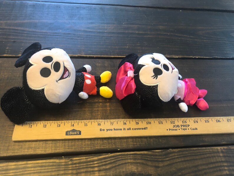 May include: Two plush toys, Mickey Mouse and Minnie Mouse, are lying on a wooden surface. Mickey is black with red shorts and yellow shoes. Minnie is black with a pink bow and pink dress. Both toys have sparkly black fabric.
