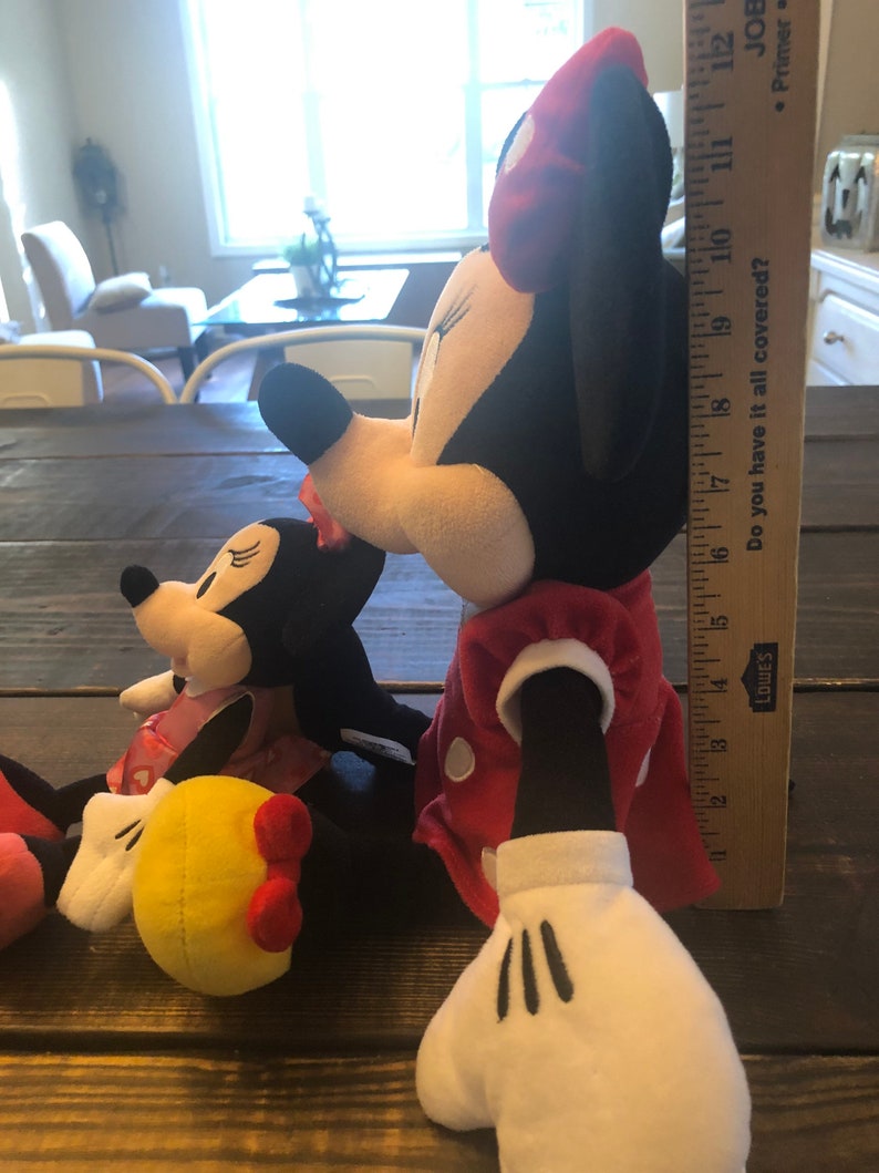 May include: Two plush Mickey Mouse toys, one red and white with black polka dots and the other black and white with red polka dots. The larger toy is holding a yellow plush ball with a red bow.