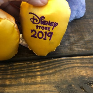 May include: A close-up of a yellow satin Disney Store tag with the text "Disney Store 2019" embroidered in purple. The tag is held by a hand, with a blurred purple plush toy in the background.