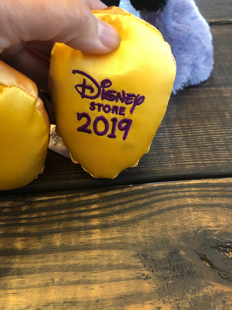 May include: A close-up of a yellow satin Disney Store tag with the text "Disney Store 2019" embroidered in purple. The tag is held by a hand, with a blurred purple plush toy in the background.