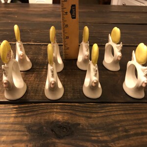 Puede incluir: Conjunto de ocho anillos de servilleta de cer&aacute;mica blanca con detalles en amarillo y rosa. Cada anillo presenta una peque&ntilde;a figura de una ni&ntilde;a con un halo amarillo.