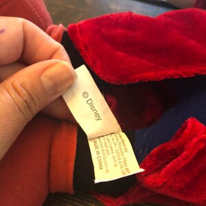 May include: A red and blue plush toy with a black collar and a white tag that says "&copy; Disney". The tag also says "New Materials: Polyester Fibers and  Fill. Reg. No. OH-22508 & PA-1190. Ages 3 years and up. For retail sale.  Amusement distribution only. MADE IN CHINA."