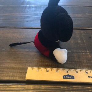 May include: A black and red plush Mickey Mouse toy sitting on a wooden surface. The toy is about 4 inches tall and has a white face and white gloves.