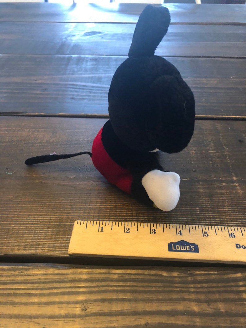 May include: A black and red plush Mickey Mouse toy sitting on a wooden surface. The toy is about 4 inches tall and has a white face and white gloves.