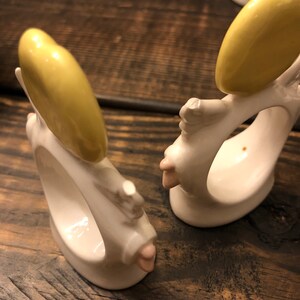 Puede incluir: Dos anillos de servilleta de cer&aacute;mica blanca con forma de &aacute;ngeles con halos amarillos y detalles en rosa. Los anillos est&aacute;n sobre una superficie de madera.