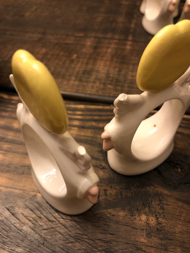 Puede incluir: Dos anillos de servilleta de cer&aacute;mica blanca con forma de &aacute;ngeles con halos amarillos y detalles en rosa. Los anillos est&aacute;n sobre una superficie de madera.