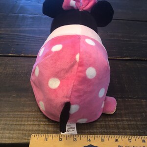 May include: Pink and white polka dot Minnie Mouse plush toy with black ears and a red bow. The toy is about 7 inches tall and has a black tail. The toy is sitting on a wooden surface with a ruler next to it. The ruler is marked with inches and has the words "Lowe's" and "Do you have it" printed on it.