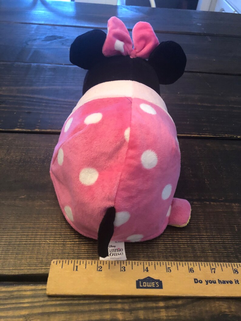 May include: Pink and white polka dot Minnie Mouse plush toy with black ears and a red bow. The toy is about 7 inches tall and has a black tail. The toy is sitting on a wooden surface with a ruler next to it. The ruler is marked with inches and has the words "Lowe's" and "Do you have it" printed on it.