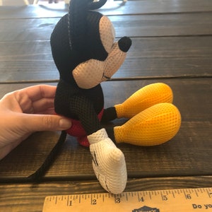 May include: A black and white mesh Mickey Mouse plush toy with yellow mesh feet. The toy is sitting on a wooden surface with a ruler next to it. The ruler is marked with inches and has the Lowe's logo on it.