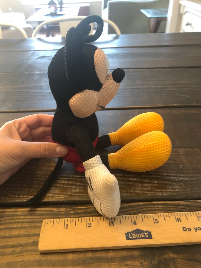 May include: A black and white mesh Mickey Mouse plush toy with yellow mesh feet. The toy is sitting on a wooden surface with a ruler next to it. The ruler is marked with inches and has the Lowe's logo on it.