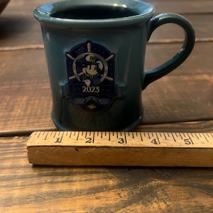 May include: Teal ceramic mug with a handle, featuring a Mickey Mouse emblem with the year 2023. The mug is approximately 3 inches tall. The mug is on a wooden surface.