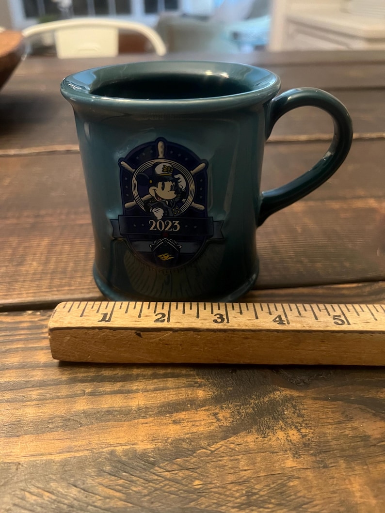 May include: Teal ceramic mug with a handle, featuring a Mickey Mouse emblem with the year 2023. The mug is approximately 3 inches tall. The mug is on a wooden surface.