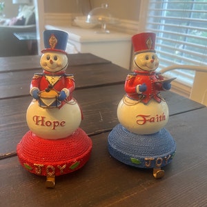 May include: Two snowman stocking holders, one in red and blue with the word "Hope" and the other in blue and white with the word "Faith". Both have a gold hook and the word "Joy" on the base.