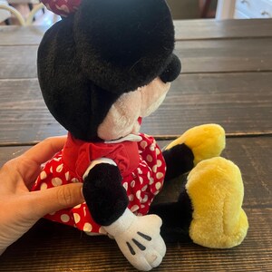 May include: A plush Minnie Mouse toy wearing a red dress with white polka dots and a red bow with white polka dots. The toy has black fur, yellow shoes, and white gloves.
