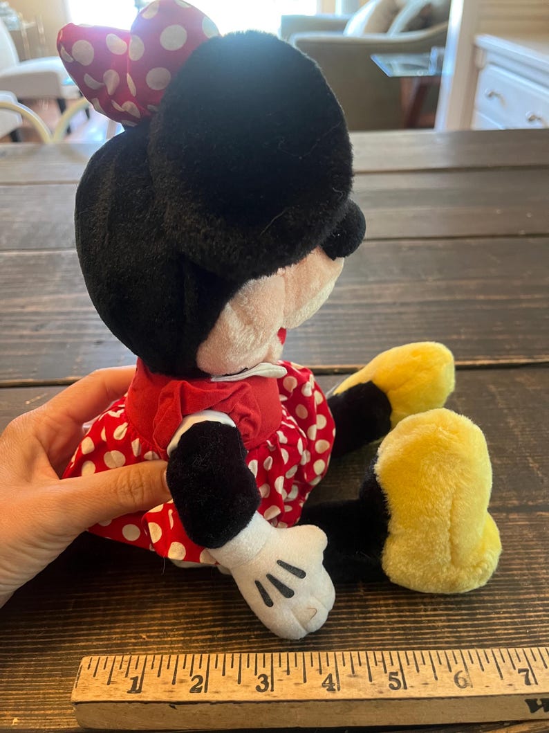 May include: A plush Minnie Mouse toy wearing a red dress with white polka dots and a red bow with white polka dots. The toy has black fur, yellow shoes, and white gloves.