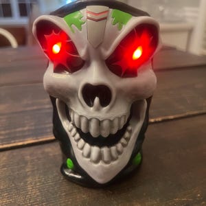 May include: A skull-shaped novelty item with glowing red eyes. The skull is primarily white with black and green accents. The mouth is open, revealing a set of teeth. The item is likely a decorative piece or a toy.