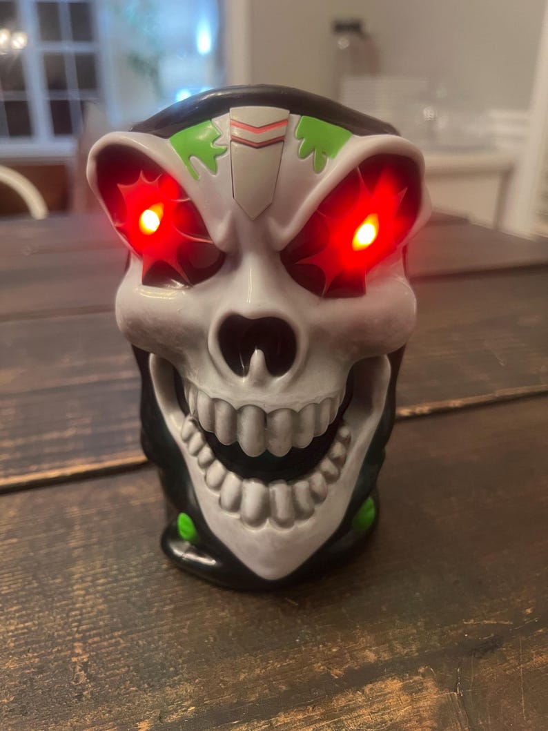 May include: A skull-shaped novelty item with glowing red eyes. The skull is primarily white with black and green accents. The mouth is open, revealing a set of teeth. The item is likely a decorative piece or a toy.