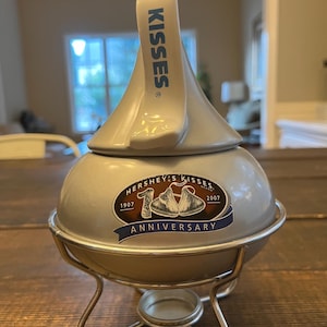 May include: A silver Hershey's Kisses candy dish with a white top that reads "KISSES". The dish has a metal stand and a sticker that says "Hershey's Kisses 100th Anniversary 1907-2007".